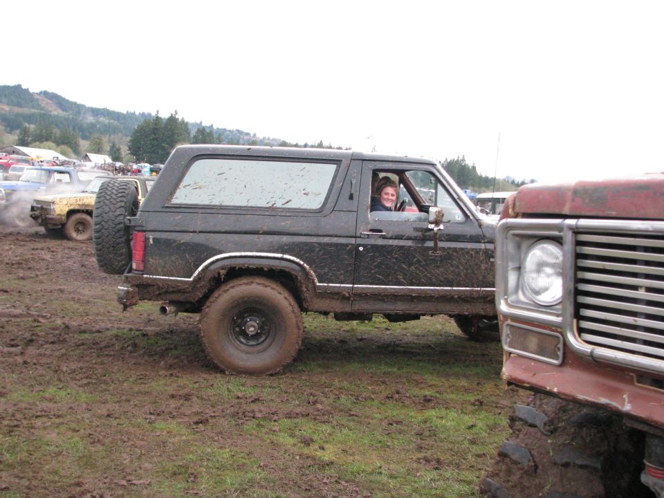 Muddy 1978 Ford Bronco with spare tire on back, driving in mud. Another vehicle is partially visible on the right