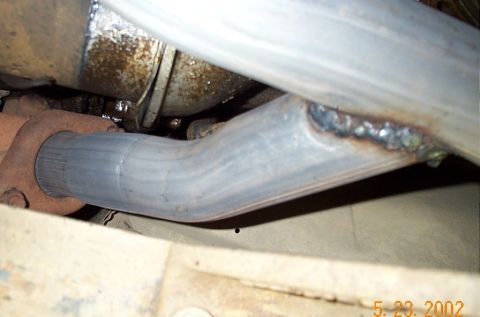 1986 Ford Bronco - Close-up underbody shot of an exhaust head pipe where a bent aluminized tube meets a larger pipe at a rough, heavily built-up weld bead. A…