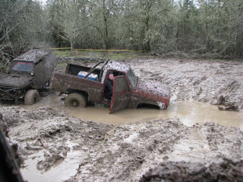Two off-road trucks stuck deep in mud, one with driver visible in open door