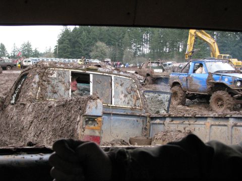 1978 Ford Bronco - Multiple off-road vehicles are heavily covered in mud during an event, with a blue pickup truck and an excavator visible in the background.
