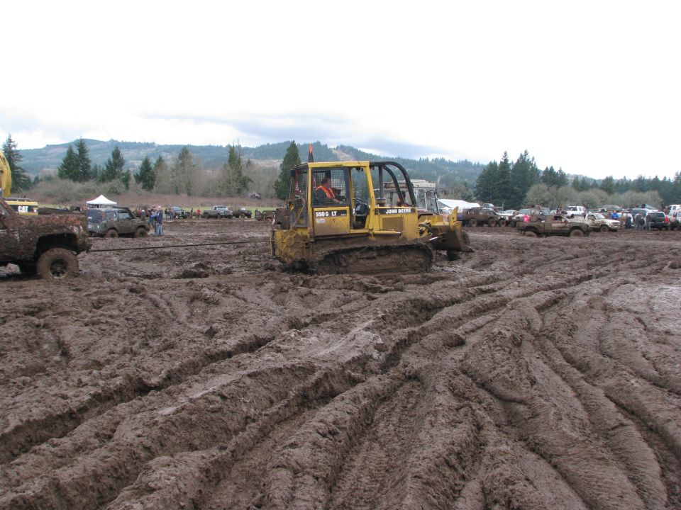 John Deere bulldozer stuck in mud, with other vehicles and trees in background