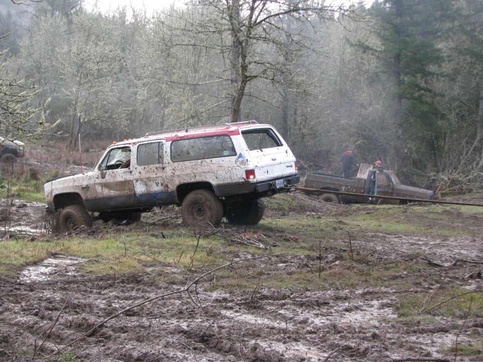 White SUV stuck in mud, being towed by brown pickup truck with two people nearby