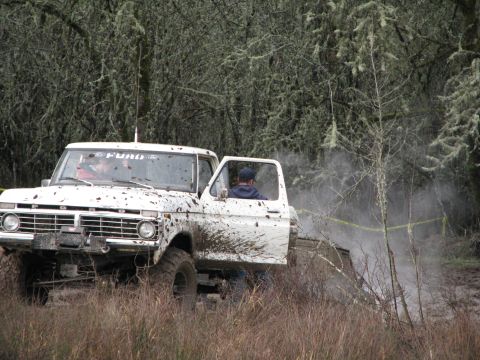 1978 Ford Bronco - A white 1973 Ford F-Series truck navigates a muddy, wooded terrain with its driver's door ajar.