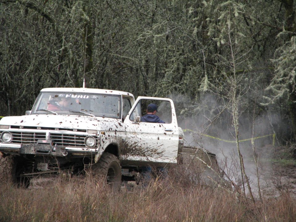 Mud-splattered white 1973 Ford F-Series truck with open driver's door, driving through a wooded area