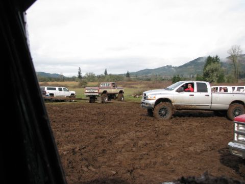 1978 Ford Bronco - Several trucks navigate a muddy field under an overcast sky, with one silver pickup truck prominently stuck.