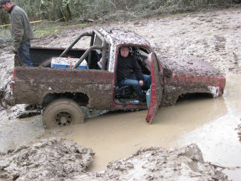 1978 Ford Bronco - A red pickup truck is deeply stuck in mud, with a person sitting in the driver's seat and another standing in the truck bed.
