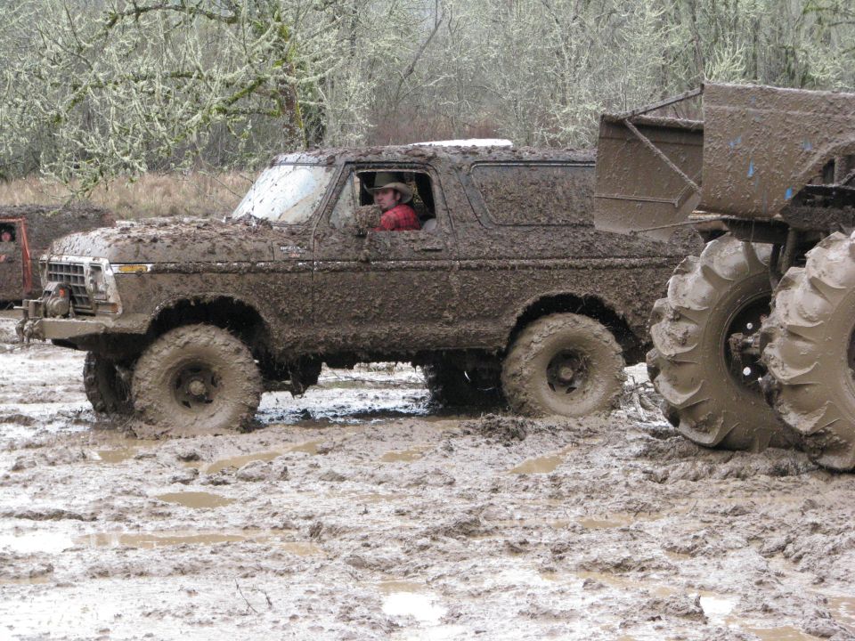 Mud-covered 1978 Ford Bronco driving through a muddy field with another vehicle partially visible