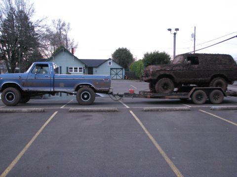 1978 Ford Bronco - A blue pickup truck tows a trailer carrying a heavily mud-caked SUV through a parking lot.
