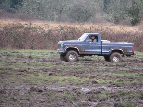 1978 Ford Bronco - A blue pickup truck navigates a muddy field under an overcast sky.