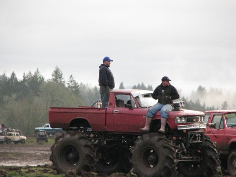 1978 Ford Bronco - Two men are on a red monster truck in a muddy area with trees in the distance.