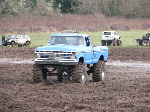 1978 Ford Bronco - A lifted blue pickup truck navigates a muddy track, with other vehicles visible in the background.