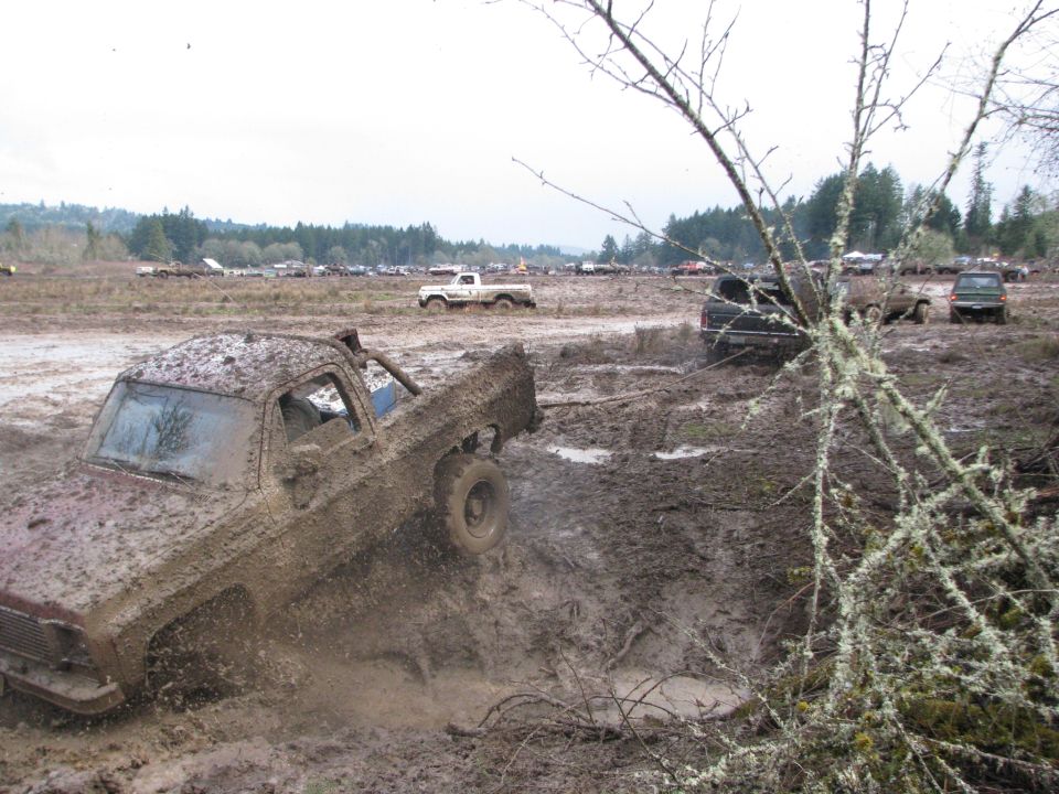 Mud-covered pickup truck struggling in a mud pit, other vehicles visible in background