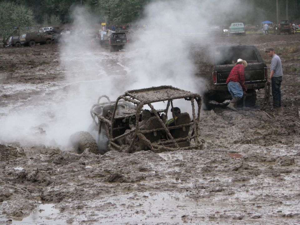Mud-covered off-road buggy stuck in mud, emitting steam, with people and other vehicles nearby