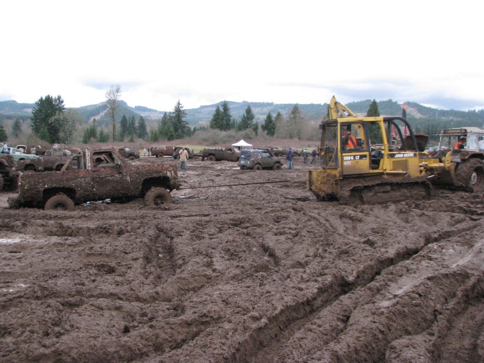 Mud-covered pickup truck being pulled by a bulldozer in a muddy field with other vehicles and trees in the background