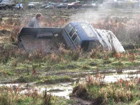 1978 Ford Bronco - A blue pickup truck is stuck deep in mud, with a person in the bed. Other vehicles are in the background.