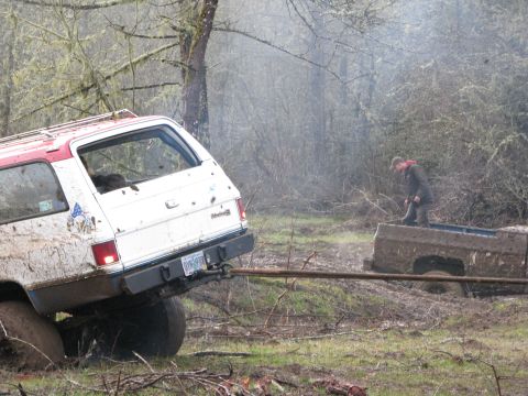 1978 Ford Bronco - A white and red SUV is stuck in the mud, being pulled by a brown truck in a wooded area.