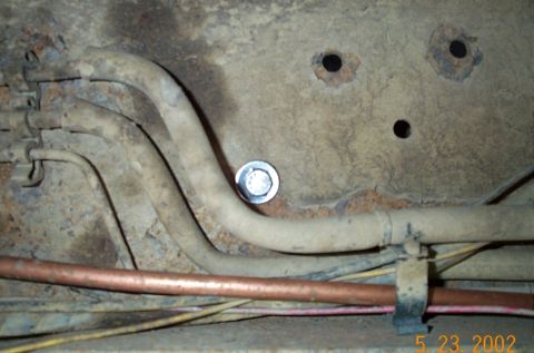 1986 Ford Bronco - Underbody detail of a dirty, surface-rusted frame rail with a bent aluminized exhaust pipe routed over it, secured near a bright new hex-he…