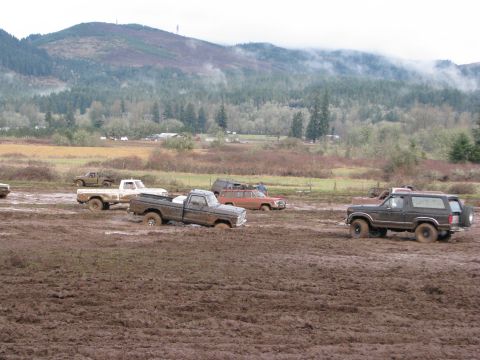 1978 Ford Bronco - Multiple off-road vehicles are stuck in a muddy field with a wooded hillside behind them.