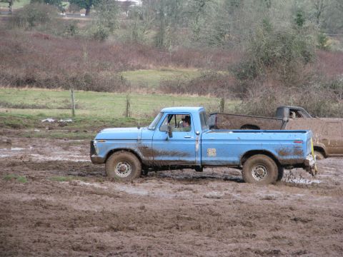 1978 Ford Bronco - A blue pickup truck navigates a muddy field, with another vehicle partially visible behind it.