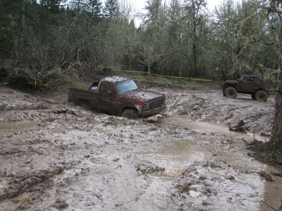 Mud-covered red pickup truck stuck in a muddy rut, with another off-road vehicle in the background