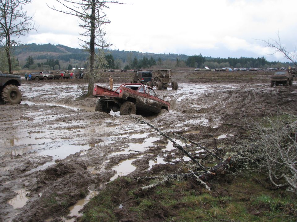 Several off-road trucks stuck in deep mud during an event, with trees and hills in the background
