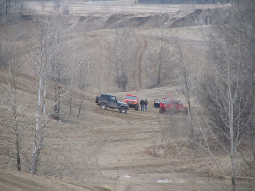 Three SUVs are on a dirt track with bare trees and hills around. One is dark gray, one is red, and one is orange
