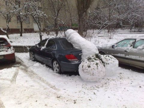 1989 Ford Bronco - dark Acura-style luxury sedan sits snow-covered in an apartment parking lot, with two large rolled snowballs piled against it.