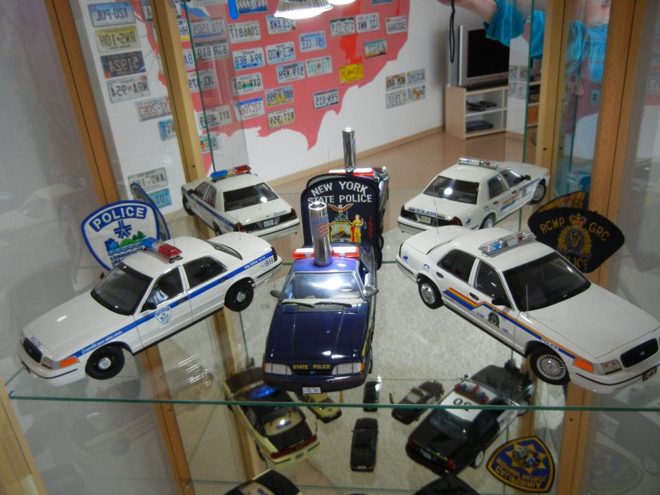 Collection of police car models, including a New York State Police car and others with police markings, displayed on glass shelves