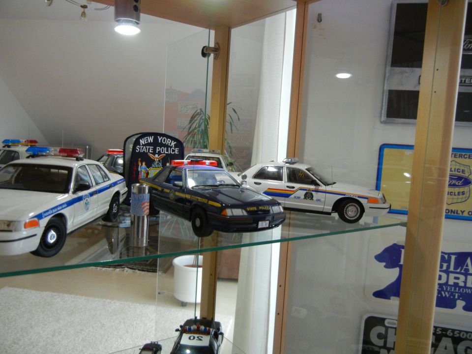 Several model police cars, including a black Ford Mustang and a white Ford Crown Victoria, are displayed in a glass cabinet with New York State Police signage