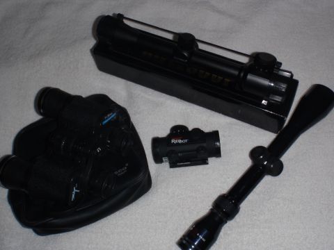 1991 Ford Bronco - A collection of optical aiming devices, including a rifle scope, red dot sight, and binoculars, are laid out on a white textured surface.