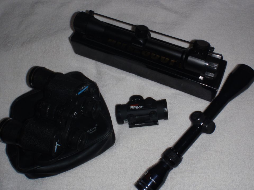 Black rifle scope, red dot sight, and binoculars on a white surface