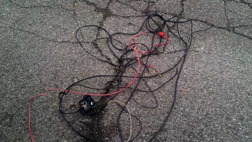 Tangled black and red electrical wires with a small black box on cracked asphalt