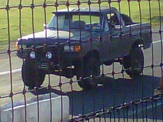 1989 Ford Bronco - A dark pickup truck is visible through a chain-link fence, a winch and an aftermarket front bumper.