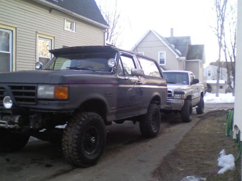 1989 Ford Bronco - A lifted, dark gray SUV with aggressive off-road tires is parked in a driveway next to a pickup truck.