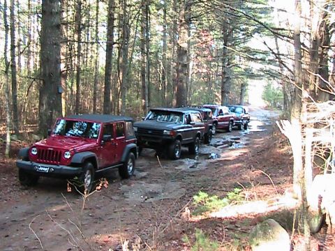 1989 Ford Bronco - A convoy of off-road vehicles navigates a wet and muddy trail through a dense forest.