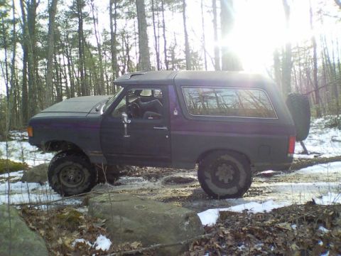 1989 Ford Bronco - 1989 Ford Bronco navigating a wooded trail with rocky obstacles and patches of snow.
