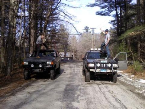 1989 Ford Bronco - Two people stand atop SUVs parked on a dirt road, surrounded by trees.