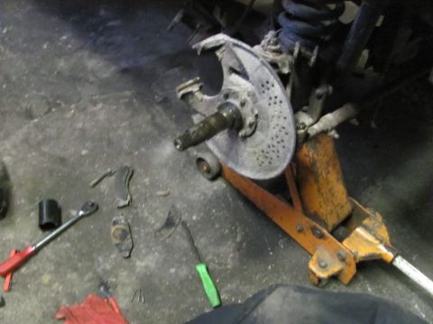 1989 Ford Bronco - Close-up of a vehicle's front wheel hub and brake rotor assembly, supported by a jack, with various tools visible on the garage floor.