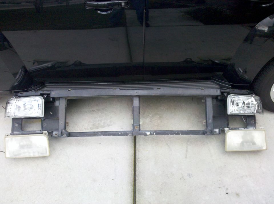 Ford truck grille shell on a driveway with clear crystal headlights beside yellowed stock ones