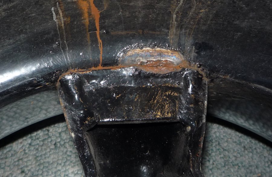 Black metal bracket with visible welds and rust streaks, on a textured surface