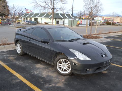 2006 Ford E350 - A dusty black seventh-generation Toyota Celica hatchback coupe shot from the front three-quarter angle in a strip-mall parking lot, with a…