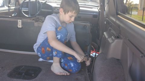 1994 Ford Bronco - A child in sports-print pajamas crouches barefoot on the gray carpeted rear floor of a Ford Bronco interior, working with pliers on wiring…