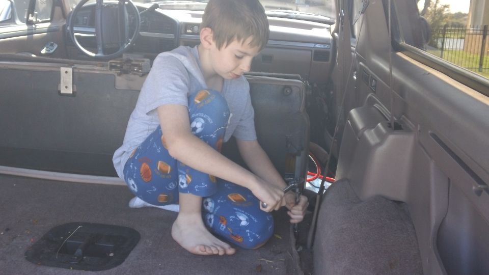 Kid working on wiring inside stripped Ford Bronco rear interior with tan dash visible