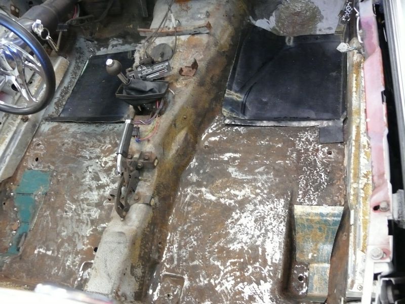 Car interior floor with rust and exposed metal, showing a manual transmission shifter and part of a steering wheel