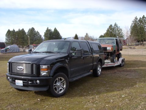 1988 Ford Bronco - A black Ford F-Series truck tows a lifted 1988 Ford Bronco on a trailer through a rural landscape.