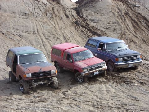 1988 Ford Bronco - Three off-road vehicles, including a blue 1988 Ford Bronco, are parked on a sandy incline.
