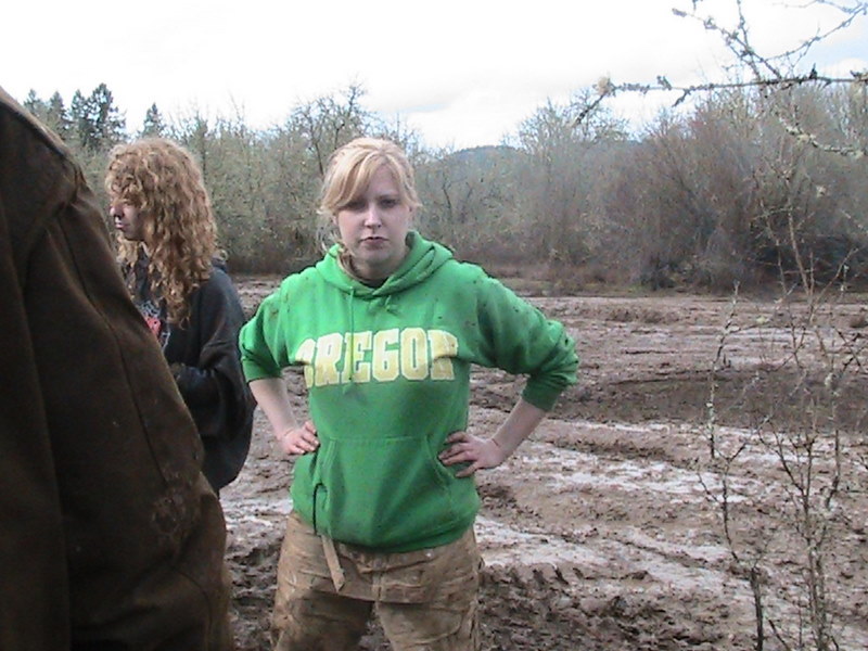 Woman in green "Oregon" hoodie and muddy pants stands in a muddy field