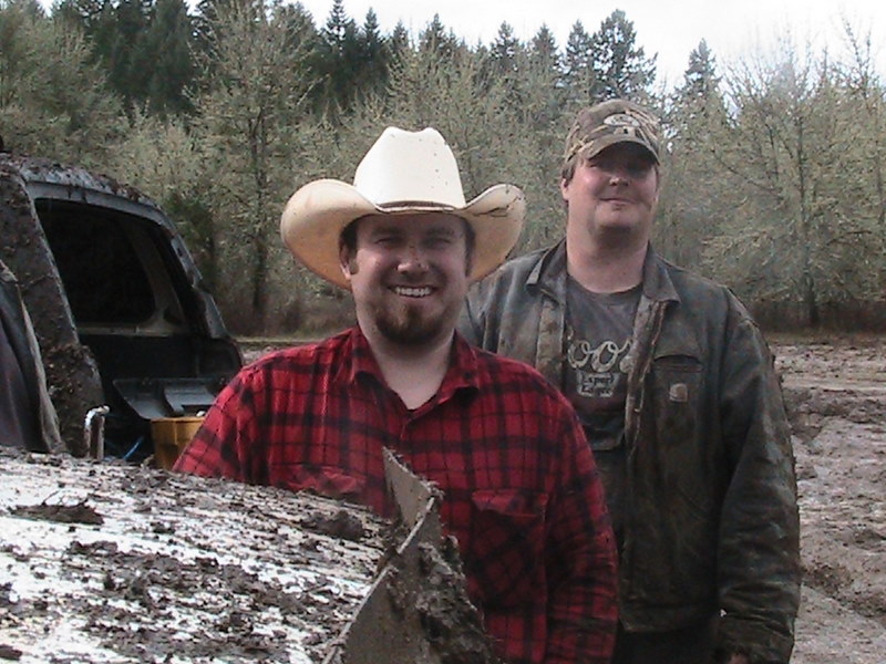 Two men, one in a cowboy hat, stand in a muddy area with trees in the background