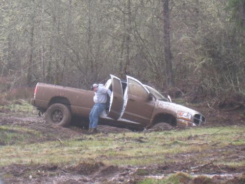 1978 Ford Bronco - A person exits a muddy pickup truck that is stuck in a wooded area.