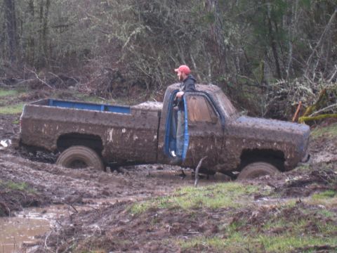 1978 Ford Bronco - A person stands in the open door of a heavily mud-covered pickup truck that is stuck in a muddy area with trees in the background.